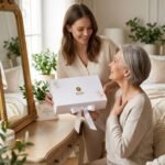 This heartwarming image shows a young woman (daughter) lovingly presenting a elegant white gift box with a ribbon to an older woman (mother/grandmother). Both are smiling warmly in a bright, cozy indoor setting. The moment captures love, gratitude, and joy, perfect for a Mother’s Day post.