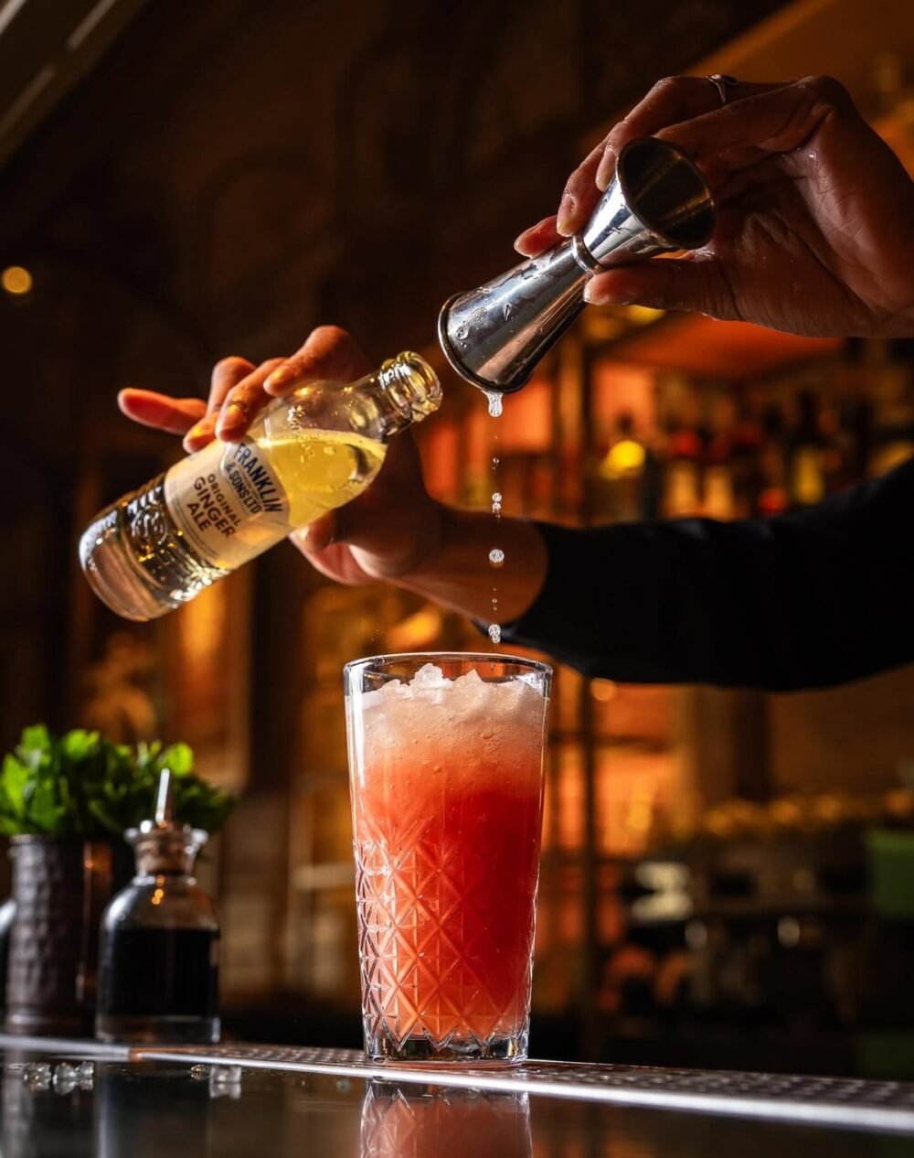 Close-up of a blender pouring a thick, vibrant red frozen strawberry daiquiri drink into a tall glass goblet garnished with a fresh lime wheel on a rustic dark wooden bar.